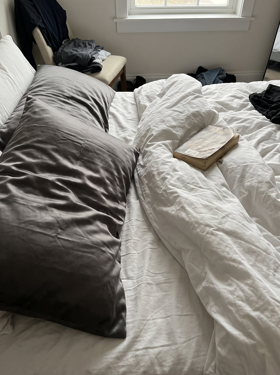 Ein dunkelgraues Seidenkissen auf einem zerwühlten Bett mit weißer Bettwäsche, daneben liegt ein Buch.