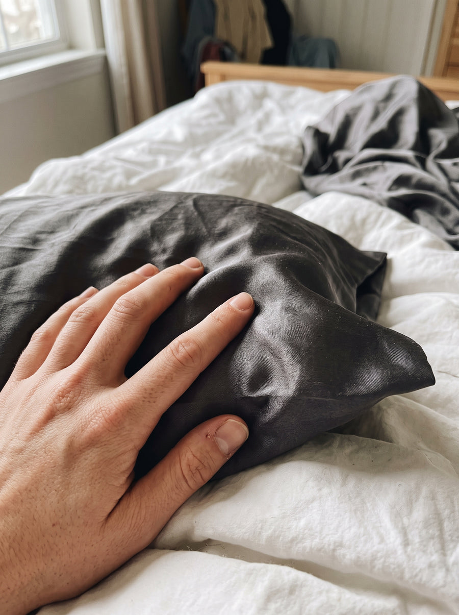 Eine Hand streicht aus der Ich-Perspektive über den glatten Stoff eines dunkelgrauen Seidenkissens auf einem Bett.